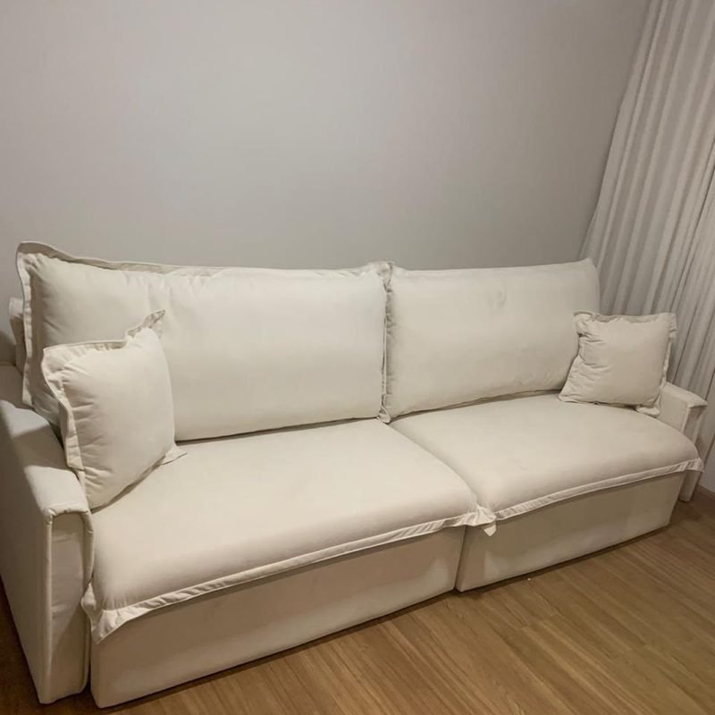 Sofá Cama Ágata em linho bege marfim, fechado, numa sala de piso de madeira com cortina clara